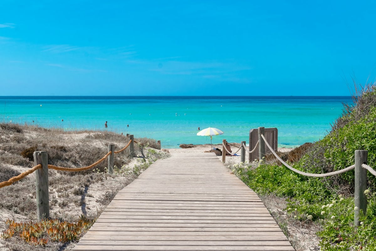 Pasarela de madera entre dunas hacia una playa de agua turquesa en Formentera